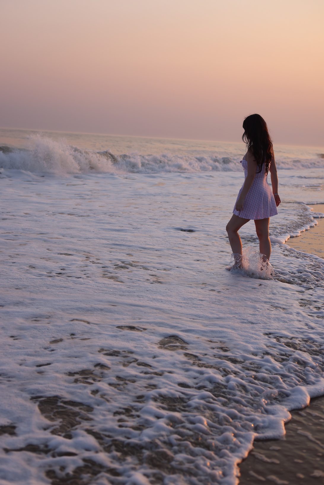 woman with long dark hair in three quarter profile wearing a pink gingham sundress standing in ocean surf at sunset