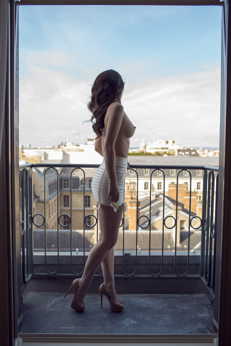 Bryleigh topless, wearing only a white slip and pink Louboutin heels, standing on a balcony gazing out at Paris