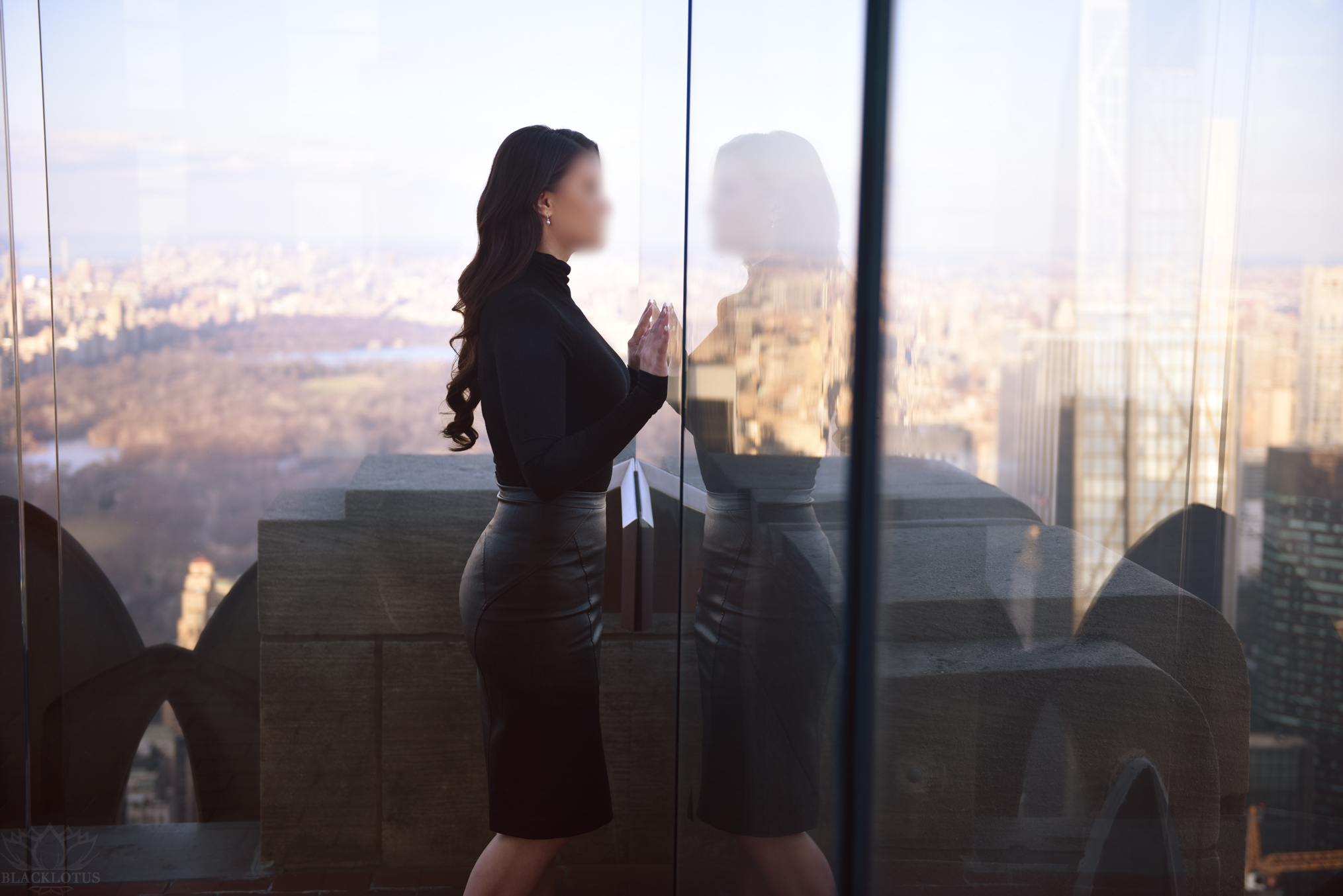 A beautiful brunette woman wearing a tight leather skirt and a black blouse stands next to a window facing her own reflection. In the distance, you can see Central Park and Manhattan.