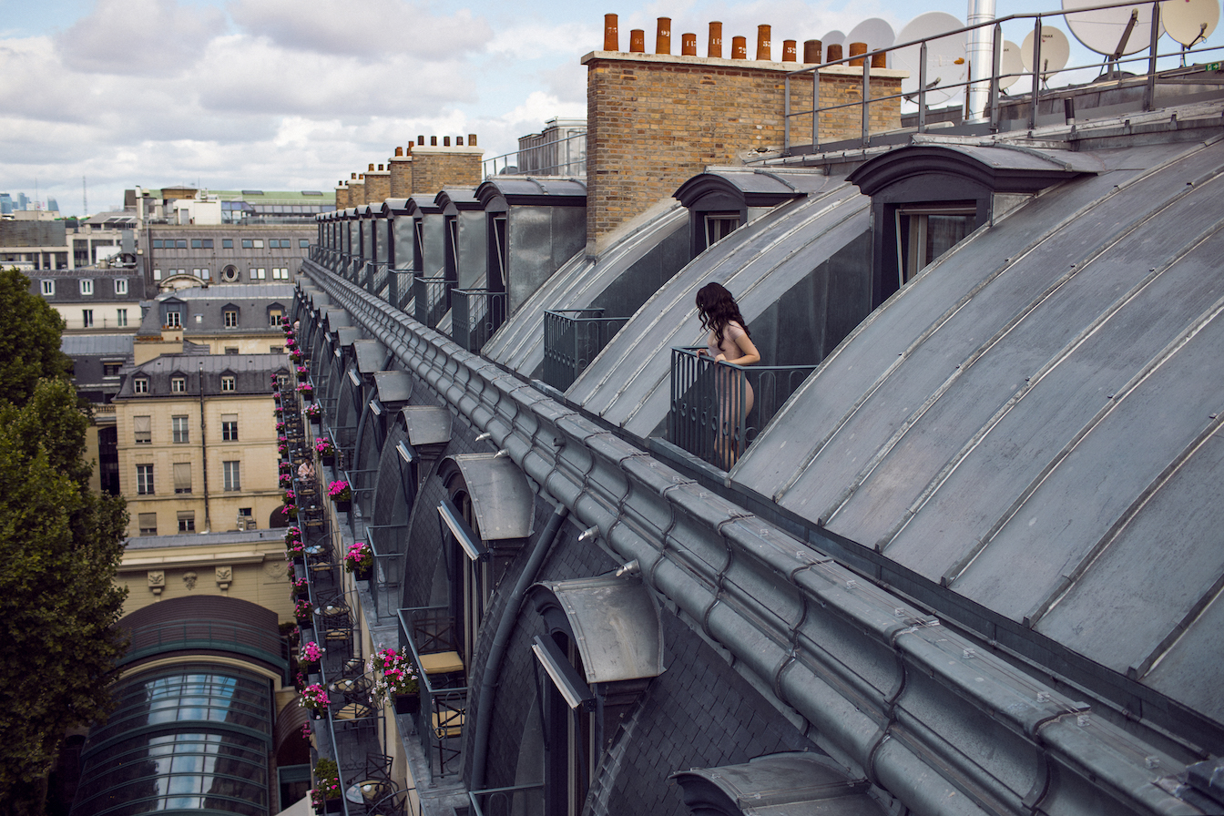 Woman on Parisian rooftop balcony overlooking zinc mansard roofs and historic architecture under cloudy skies