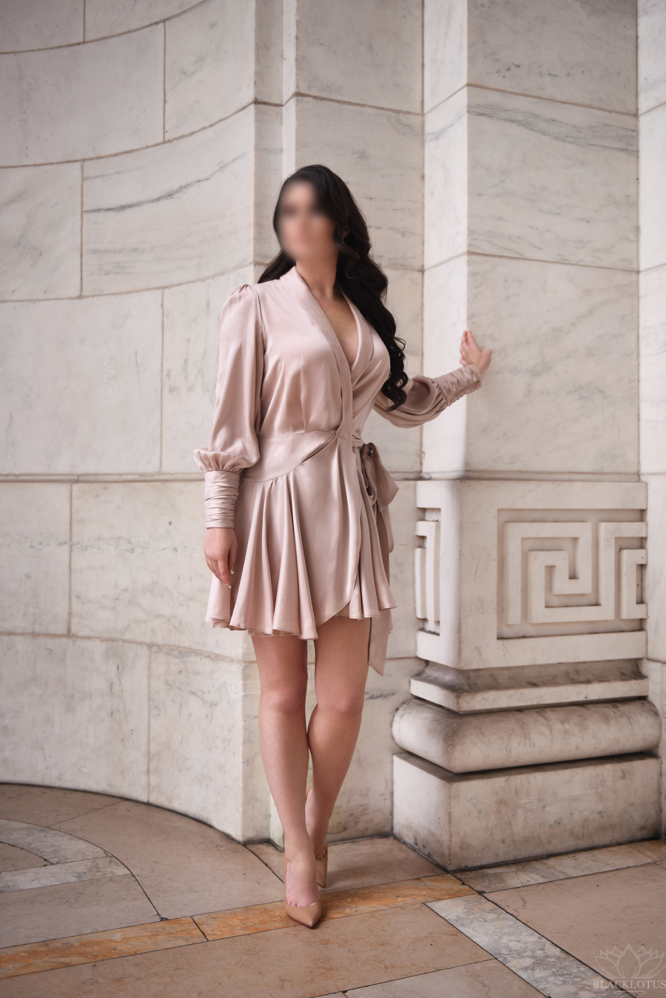 Woman in elegant champagne wrap dress posing by ornate marble column architecture in natural light.