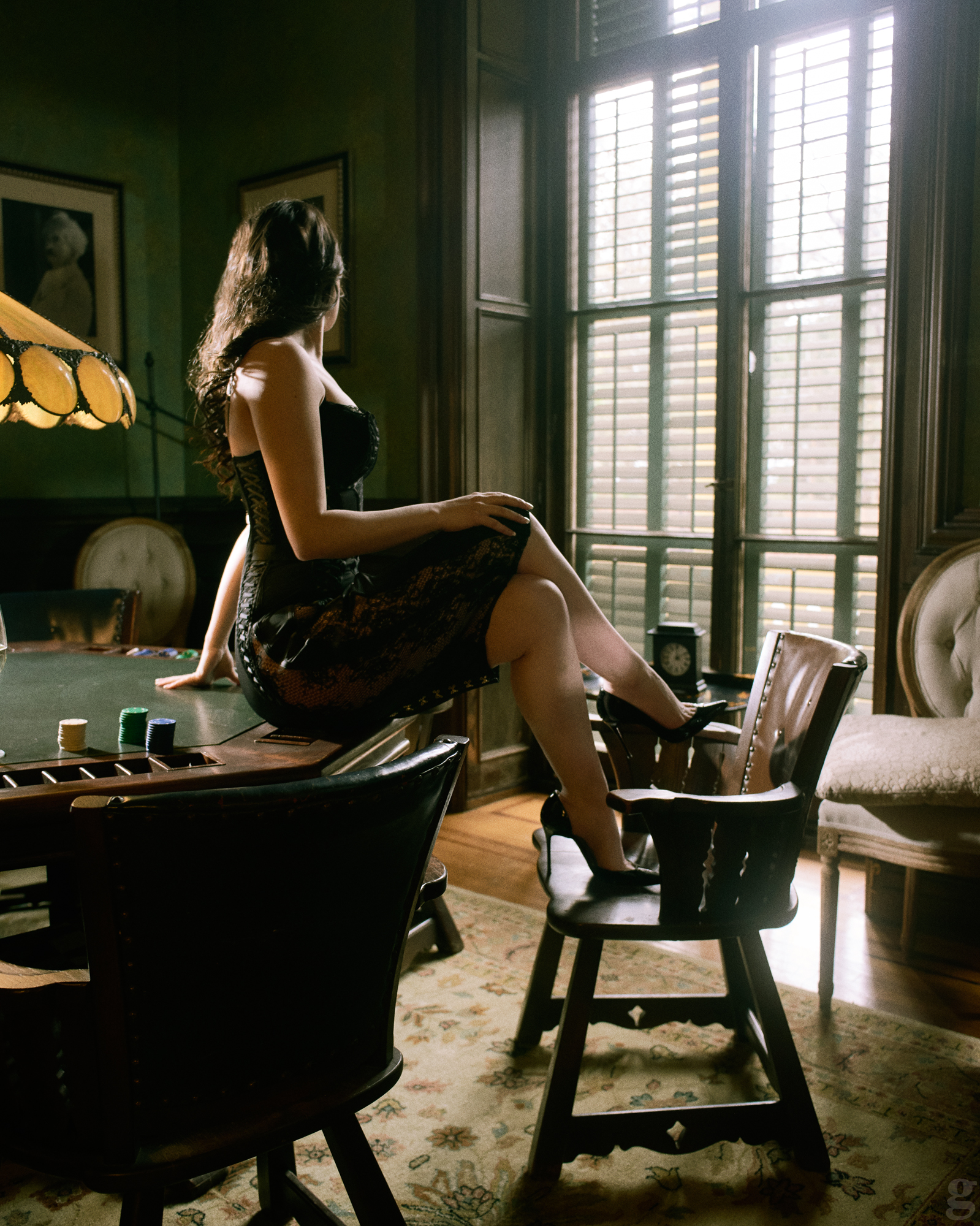 Woman in black dress sitting on desk by sunlit window shutters in elegant vintage room