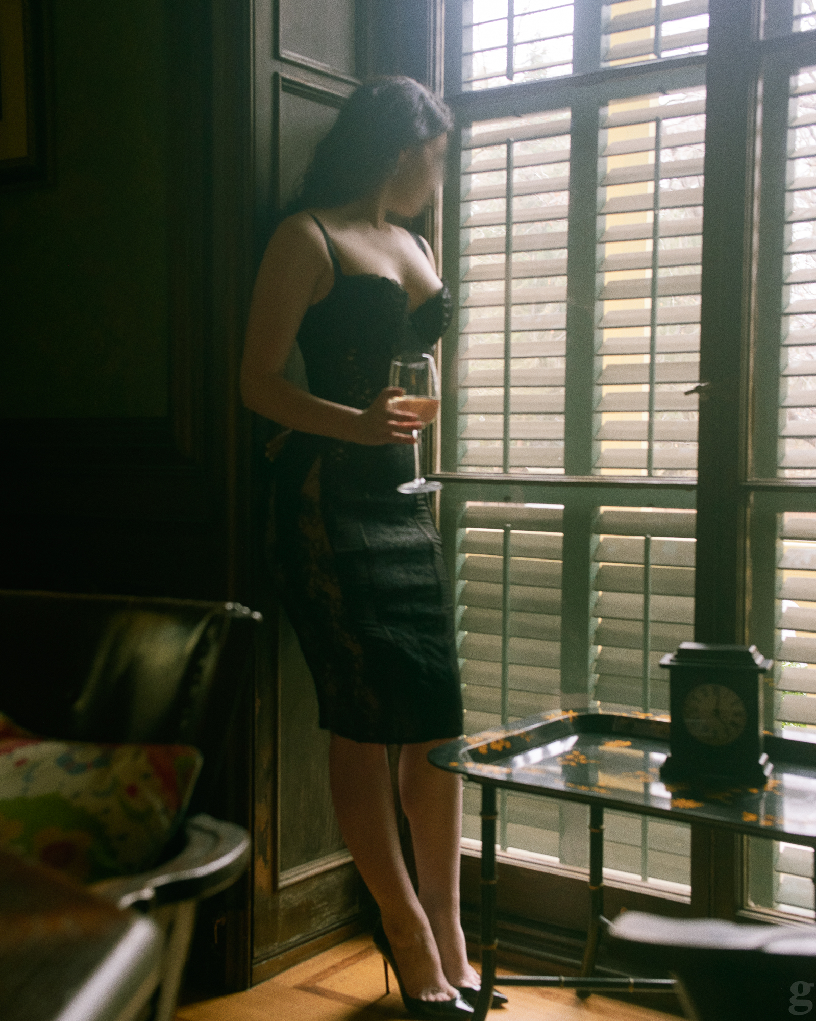 Woman in black dress holding wine glass by window with shutters in moody, film-inspired interior setting