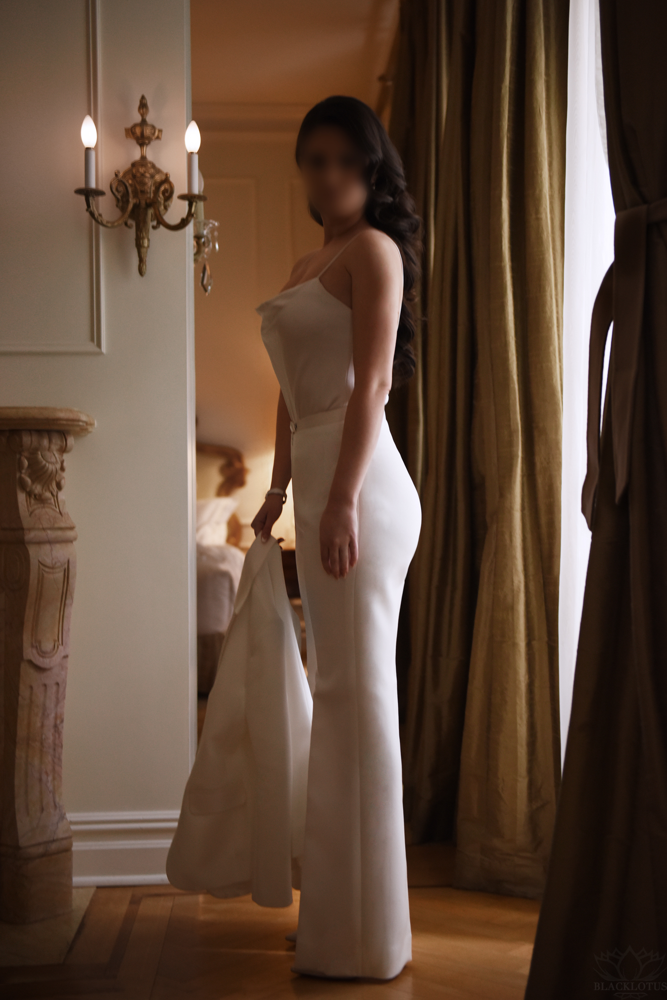 Woman in elegant white gown standing in luxurious room with ornate wall sconce and golden drapes