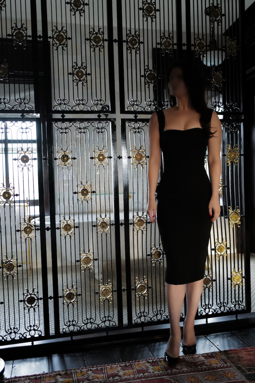 Woman in elegant black dress standing before ornate decorative iron gates with golden medallion details in natural light.