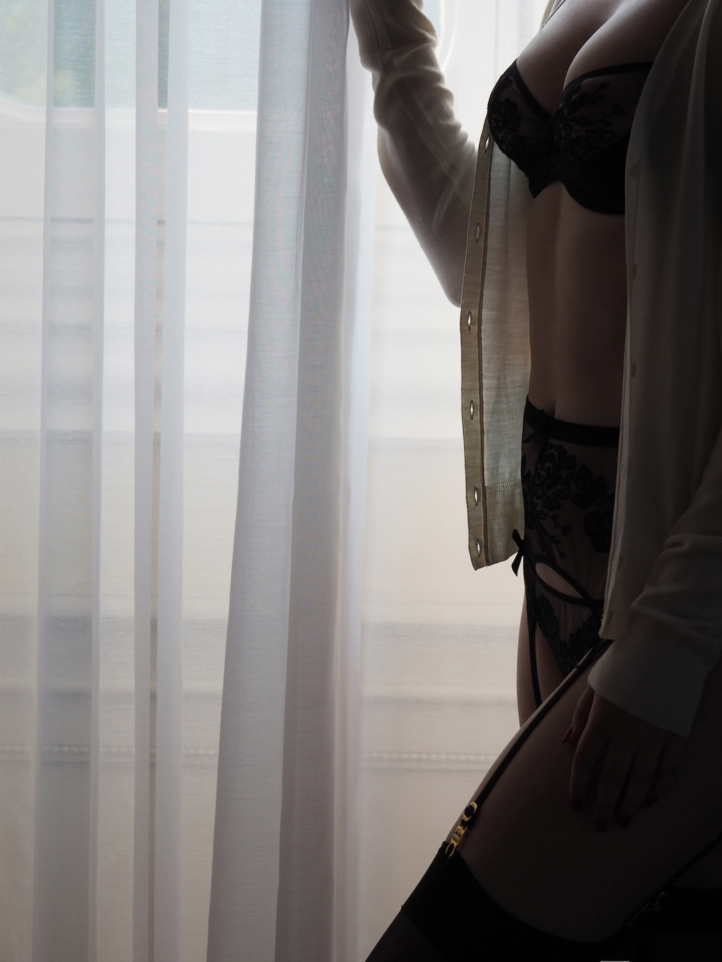 Woman in elegant lingerie and unbuttoned shirt standing by sheer curtains overlooking serene waterfront at dawn