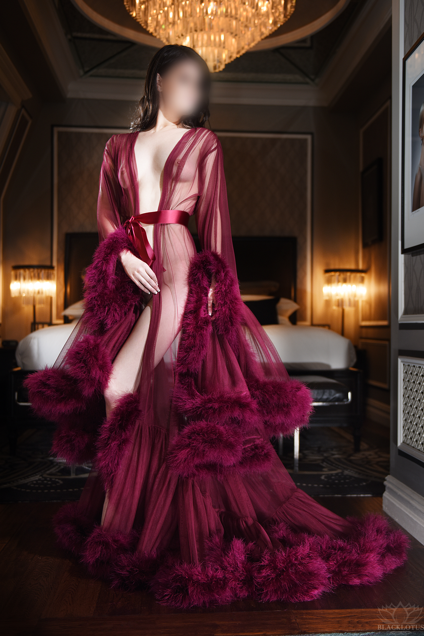 Woman in burgundy feathered robe standing in elegant hotel suite with crystal chandelier overhead