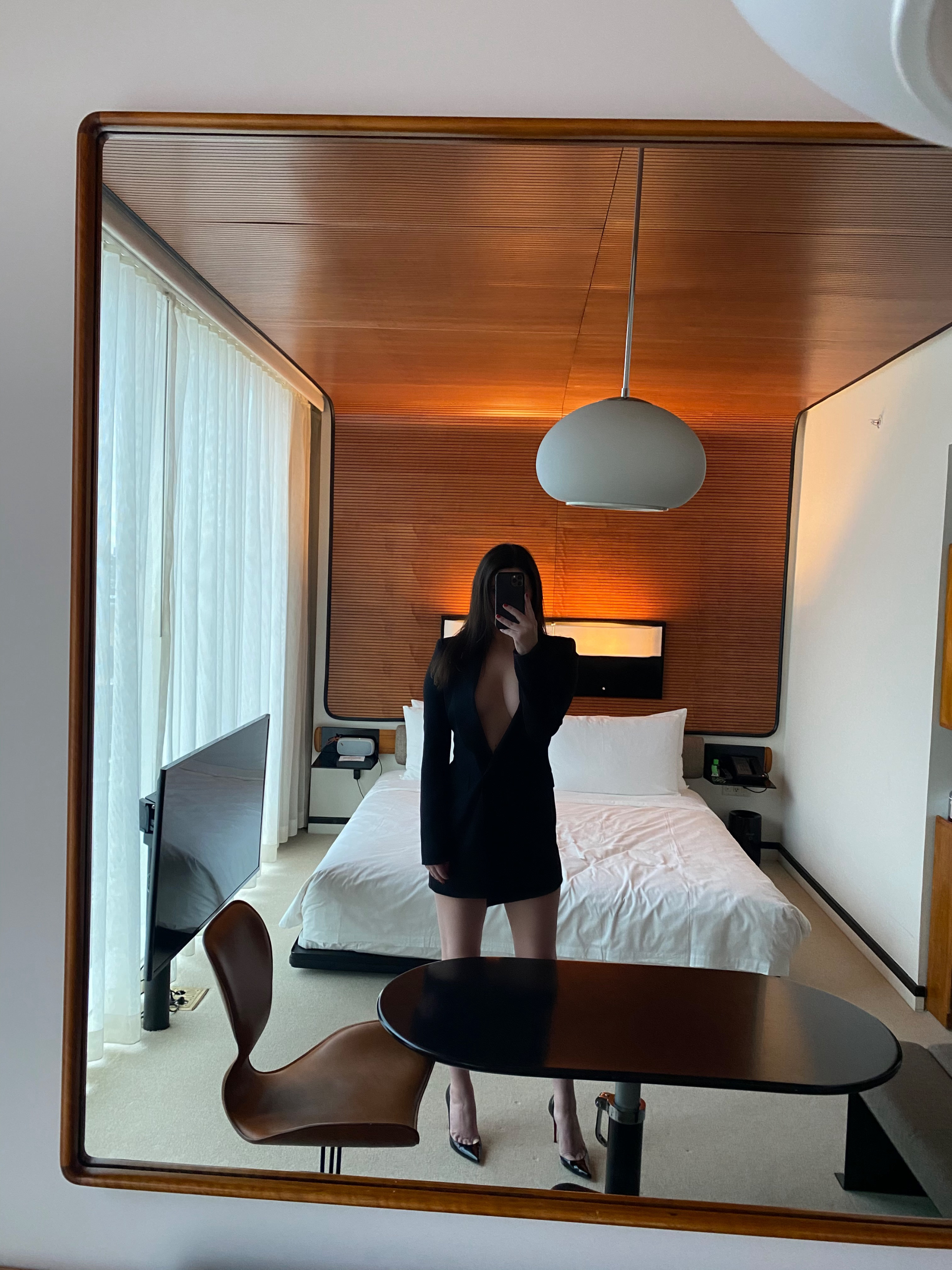 Woman in black dress taking mirror selfie in modern hotel room with warm wood ceiling and minimalist design
