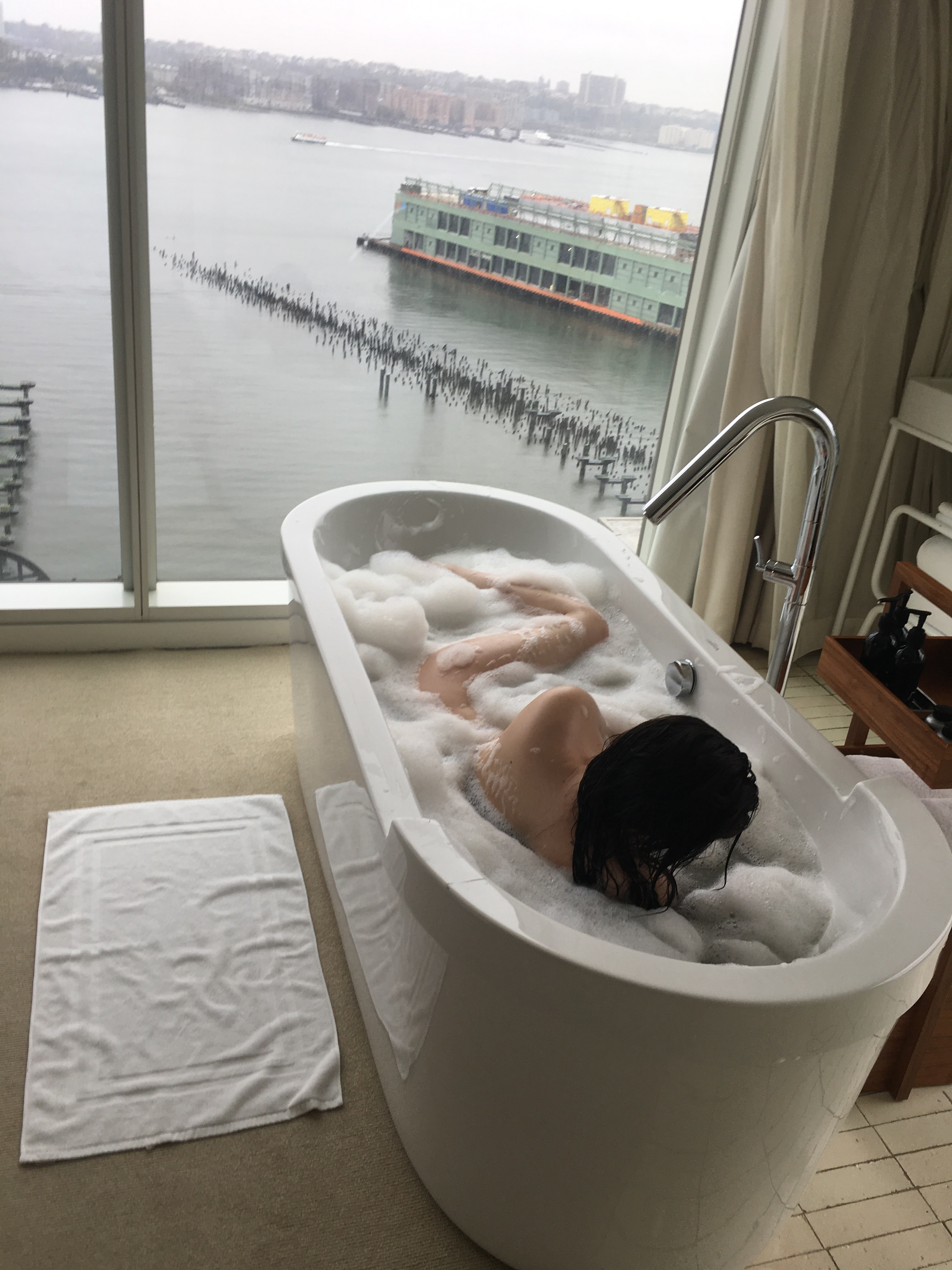 Person relaxing in freestanding bathtub with river and ferry boat view through floor-to-ceiling windows