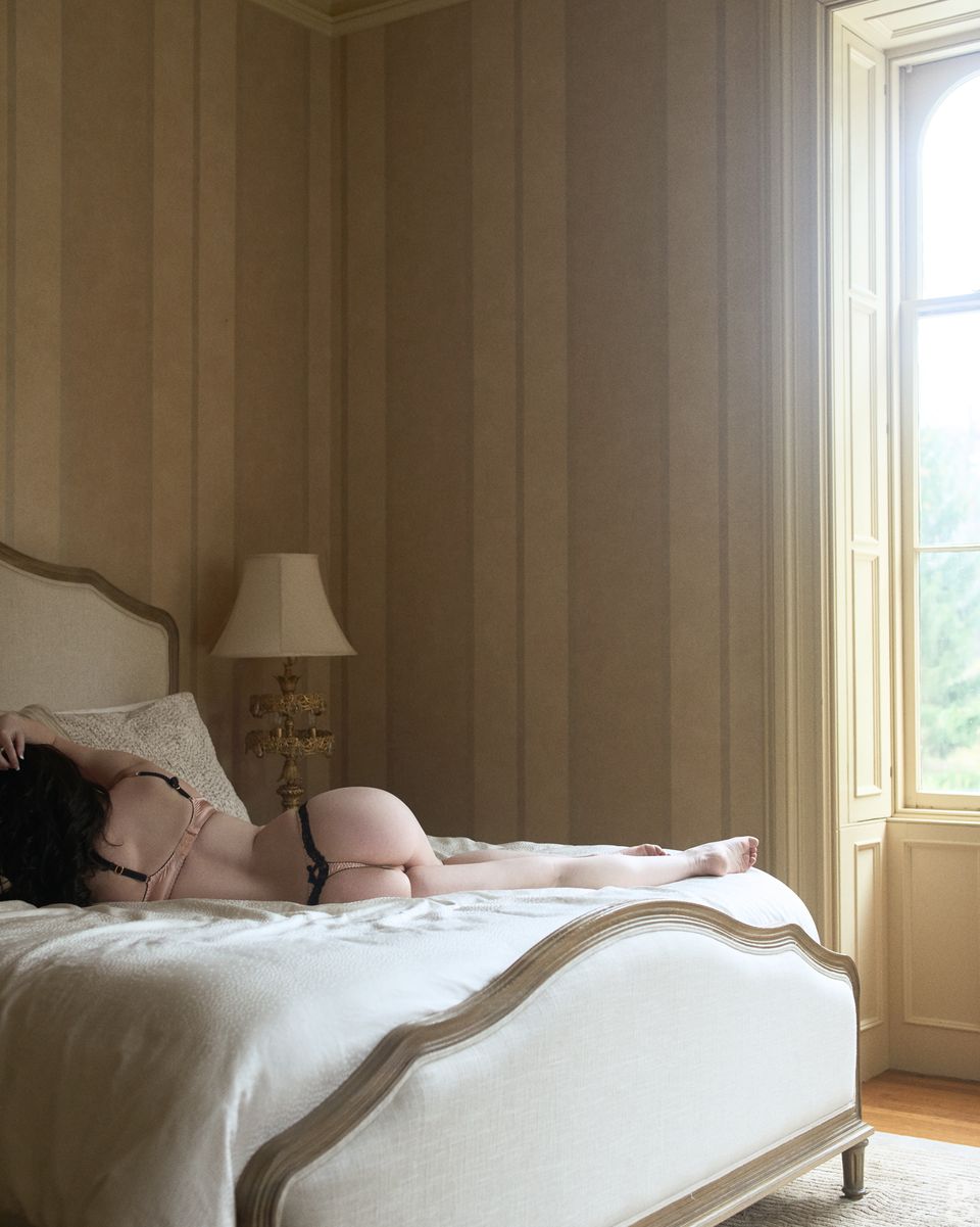 Woman reclining on elegant upholstered bed in sunlit room with striped wallpaper and classic French doors