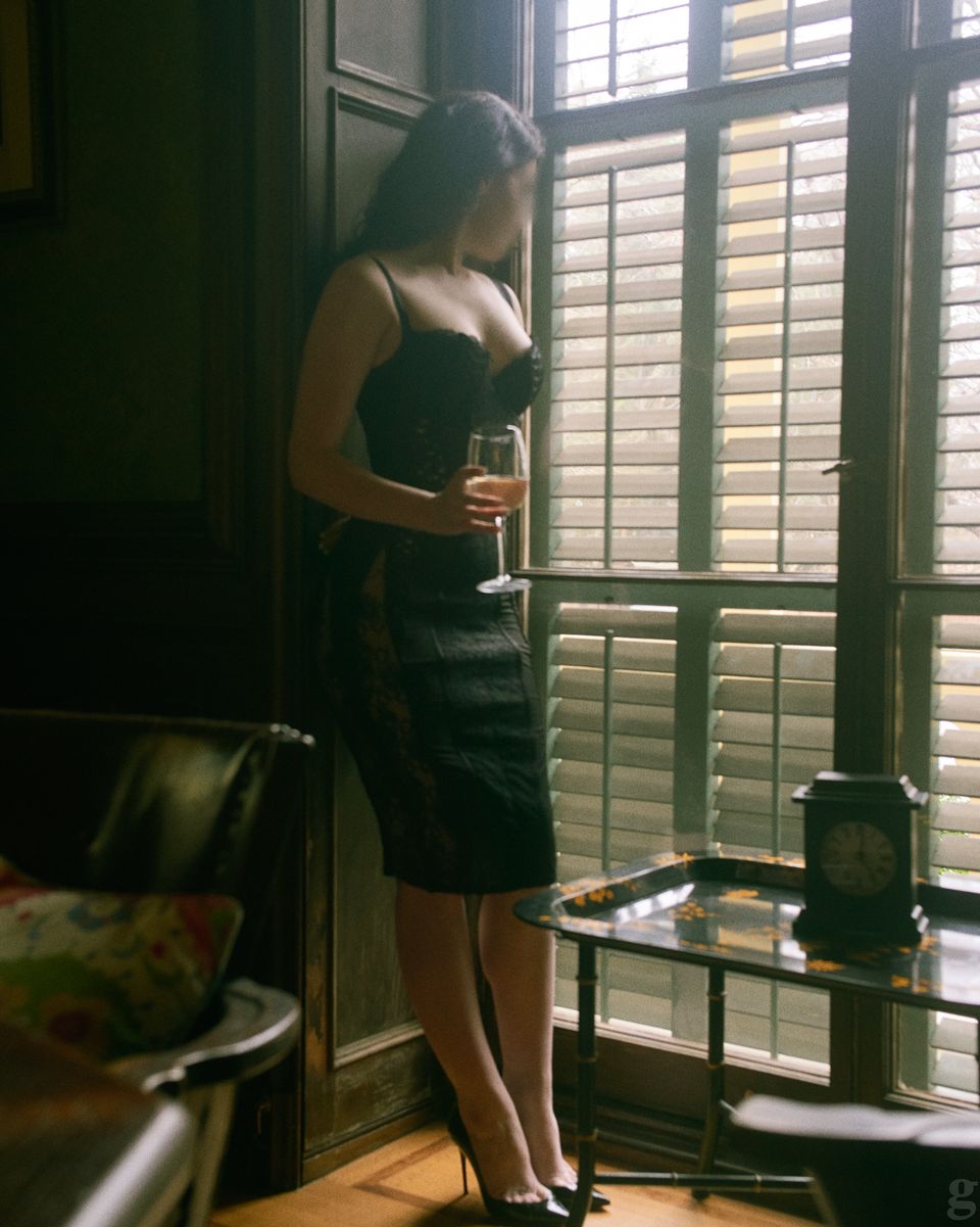 Woman in black dress holding wine glass by window with shutters in moody, film-inspired interior setting