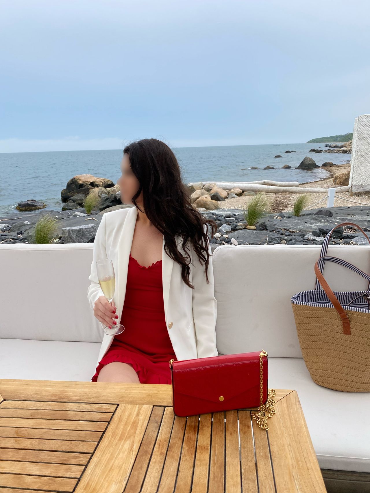 Woman in red dress and white blazer enjoying champagne at a seaside terrace overlooking rocky coastal waters